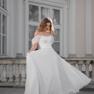 May include: A woman in a white off-the-shoulder wedding dress with a fitted bodice and a flowing skirt. She is standing in front of a white building with large windows.