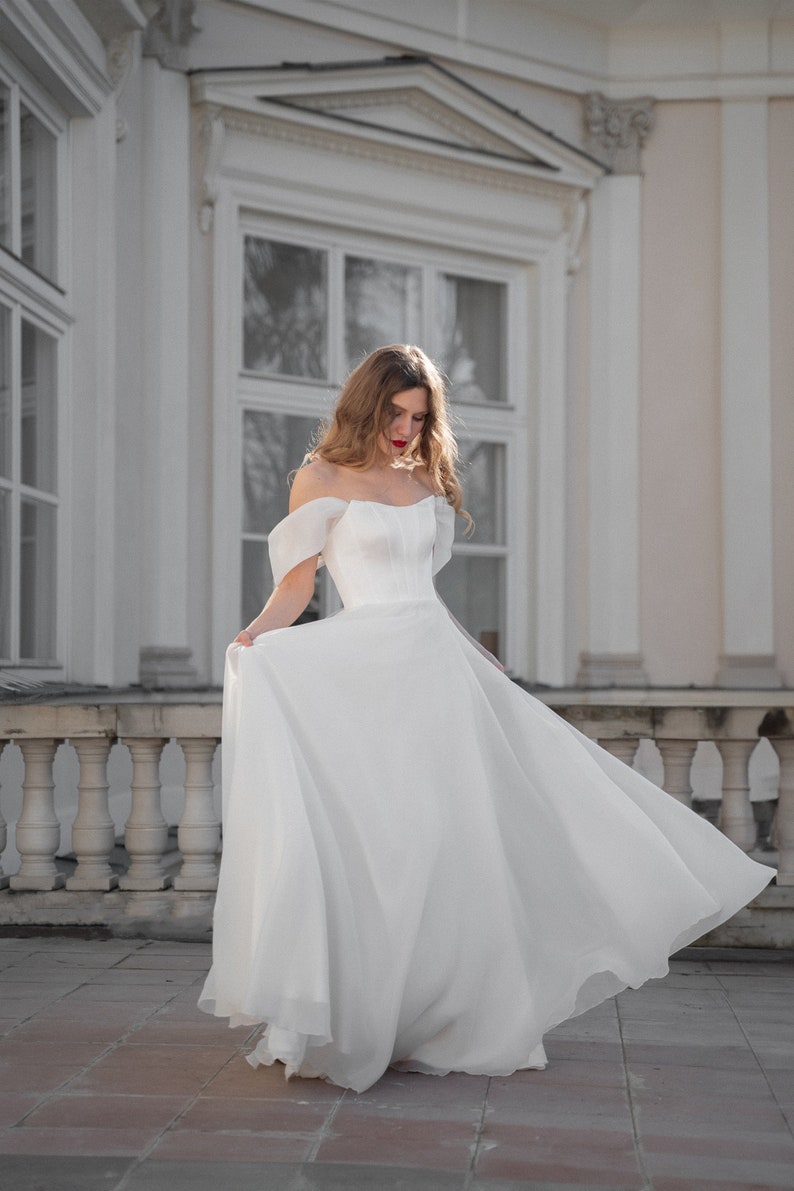 May include: A woman in a white off-the-shoulder wedding dress with a fitted bodice and a flowing skirt. She is standing in front of a white building with large windows.