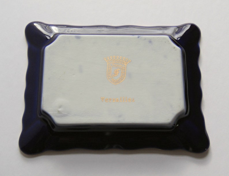 May include: A rectangular, ceramic tray with a wavy, dark blue border. The center is white with a gold emblem and the word "Versailles" in gold lettering. The tray is likely for decorative use.