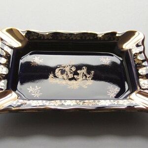 May include: A rectangular ashtray with a navy blue interior and gold trim. The ashtray features a gold illustration of figures in a garden scene. The edges are scalloped and decorated with gold floral patterns.