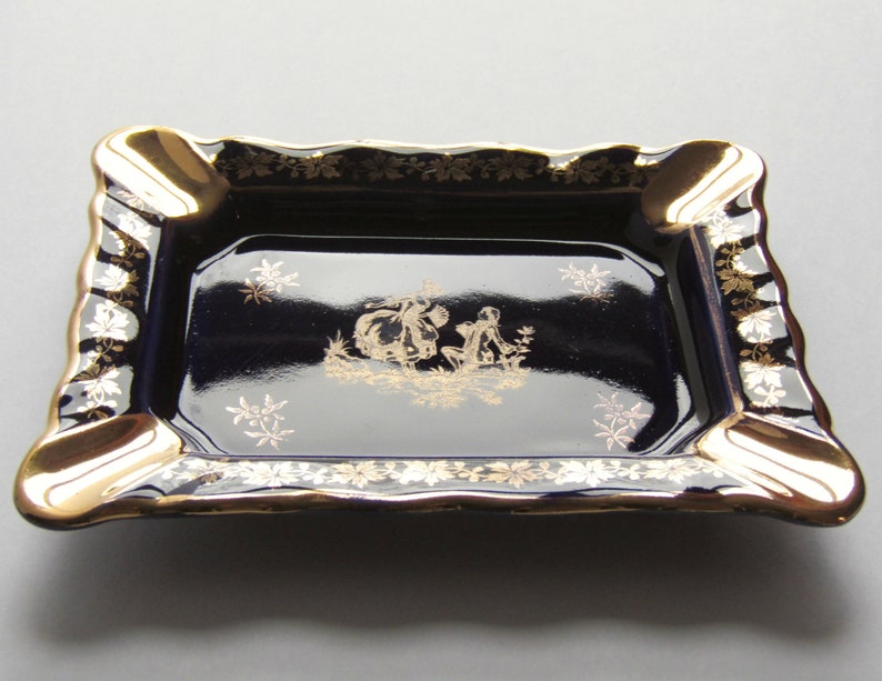 May include: A rectangular ashtray with a navy blue interior and gold trim. The ashtray features a gold illustration of figures in a garden scene. The edges are scalloped and decorated with gold floral patterns.