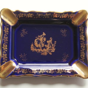 May include: A rectangular ashtray with a deep blue base and gold accents. The ashtray features a gold rim, four cigarette rests, and a gold illustration of figures. The edges are scalloped, and the design includes gold floral patterns.