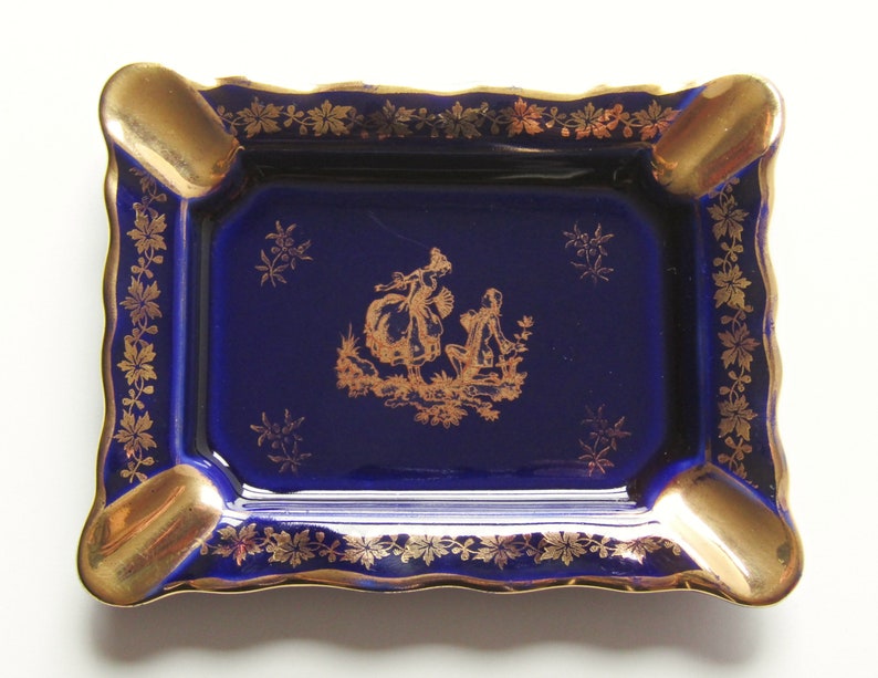 May include: A rectangular ashtray with a deep blue base and gold accents. The ashtray features a gold rim, four cigarette rests, and a gold illustration of figures. The edges are scalloped, and the design includes gold floral patterns.