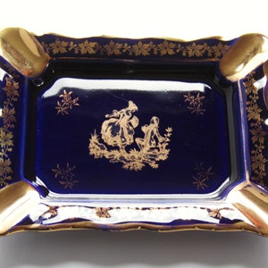 May include: Rectangular ashtray with a deep blue interior and gold trim. The ashtray features a gold floral border and four gold resting areas. A gold illustration of two figures is in the center of the ashtray.
