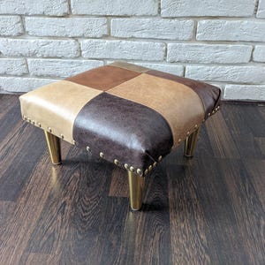 May include: A square-shaped, patchwork ottoman with a brown, tan, and beige leather upholstery. The ottoman has four gold-colored legs and is sitting on a dark wood floor.
