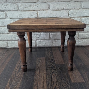 May include: A small, square wooden table with a dark brown finish. The tabletop has a parquet design, and the legs are turned and rounded. The table is set against a white brick wall and a dark wood floor.