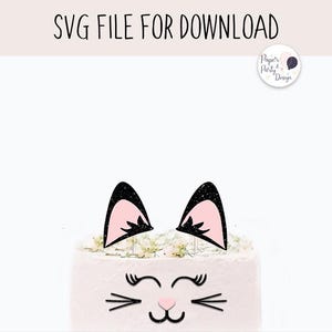 May include: A white cake with a pink and black cat face design. The cat has black ears, pink inside the ears, black whiskers, and pink eyes.