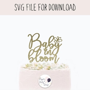 May include: A gold glitter cake topper with the words "Baby in Bloom" in a script font, topped with a flower design. The cake topper is on a white frosted cake with small white flowers. The text "SVG FILE FOR DOWNLOAD" is at the top.