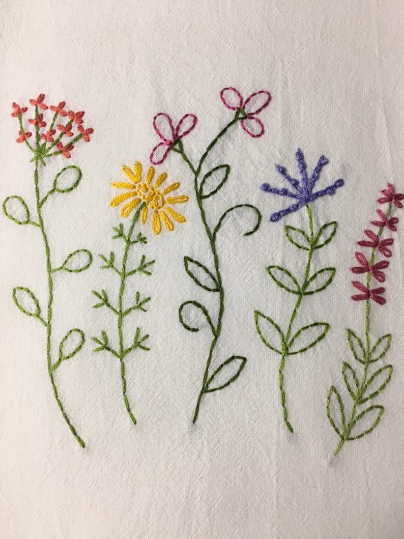Puede incluir: Dise&ntilde;o floral bordado con cinco tipos diferentes de flores en tonos rojos, amarillos, morados y rosas. Las flores tienen tallos y hojas verdes, bordadas sobre un fondo de tela blanca.
