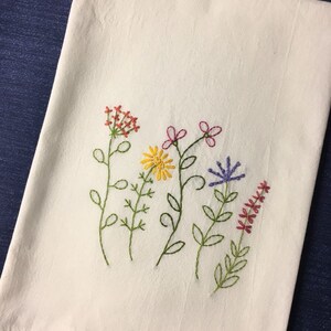 Puede incluir: Un pa&ntilde;o de cocina de lino blanco bordado con flores silvestres de colores. El bordado presenta flores rojas, amarillas, rosas y moradas con tallos y hojas verdes. El pa&ntilde;o est&aacute; doblado y expuesto sobre una superficie azul.