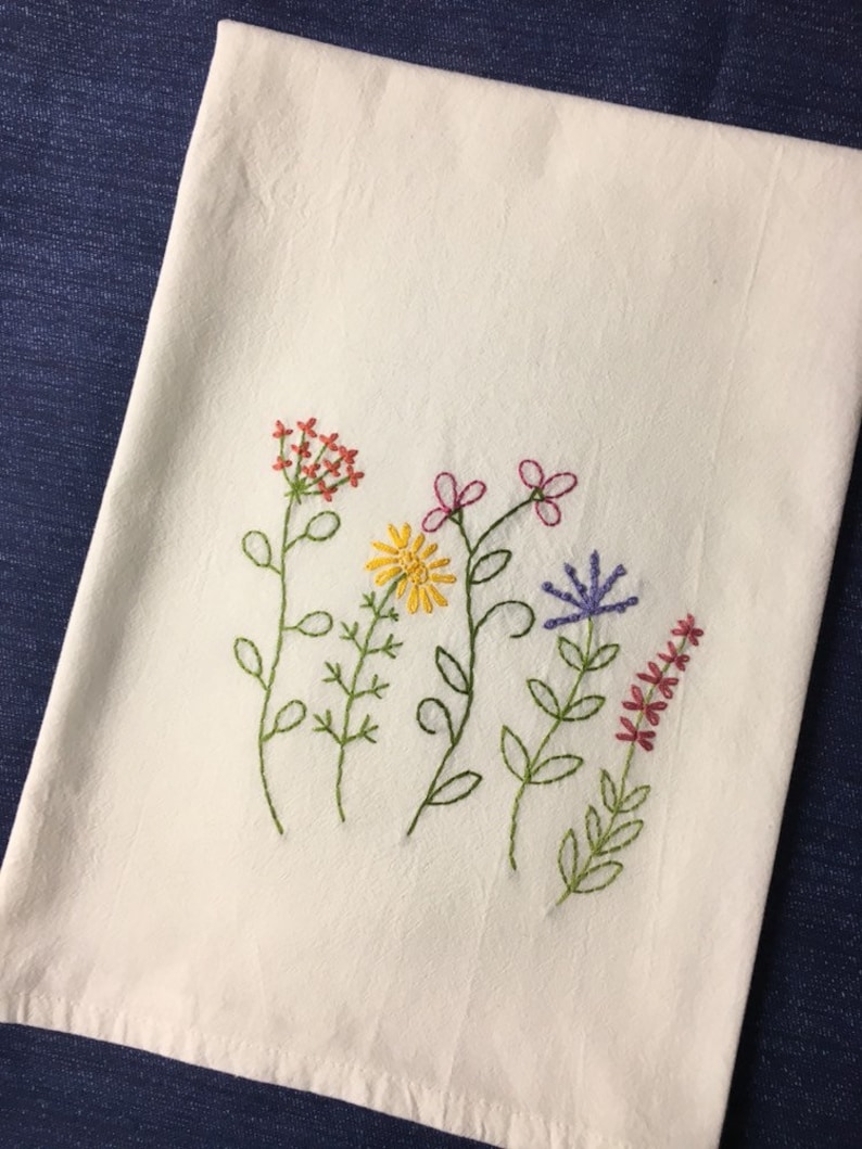 Puede incluir: Un pa&ntilde;o de cocina de lino blanco bordado con flores silvestres de colores. El bordado presenta flores rojas, amarillas, rosas y moradas con tallos y hojas verdes. El pa&ntilde;o est&aacute; doblado y expuesto sobre una superficie azul.
