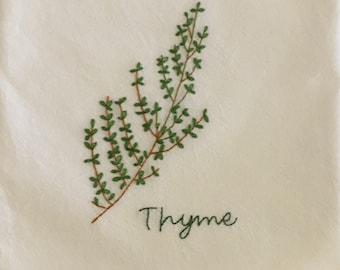 May include: Embroidered sprig of thyme on a cream-colored fabric. The thyme is depicted with green leaves and a brown stem. The word "Thyme" is embroidered in green script below the sprig.