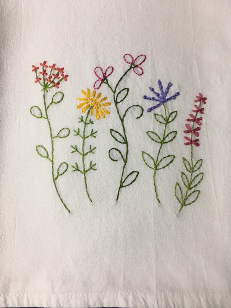 Puede incluir: Tela blanca con bordados de flores silvestres en tonos rojos, amarillos, morados y rosas. Las flores est&aacute;n bordadas con tallos y hojas verdes. El bordado es un dise&ntilde;o floral decorativo.