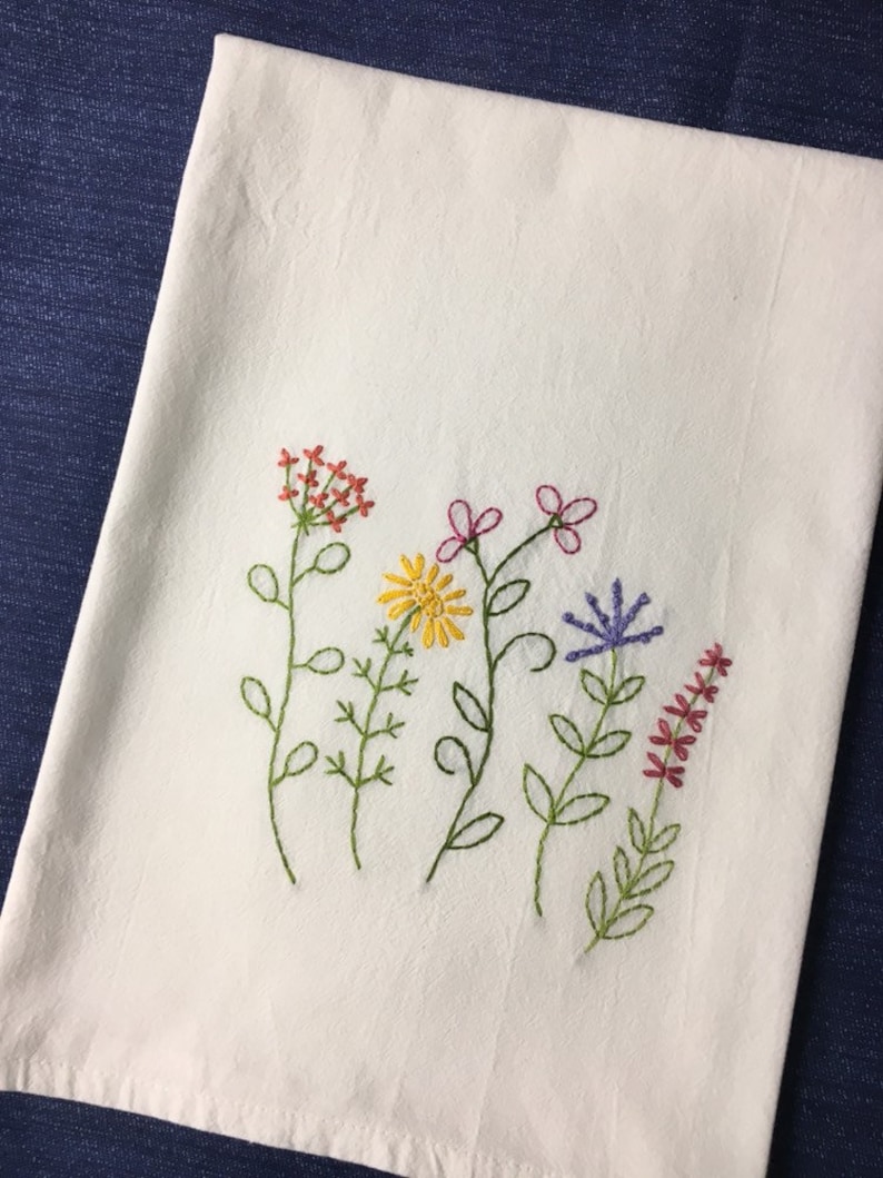 Puede incluir: Pa&ntilde;o de cocina blanco con bordados de flores silvestres en rojo, amarillo, rosa y morado. Las flores est&aacute;n bordadas con tallos y hojas verdes. El pa&ntilde;o es de tela suave y texturizada.