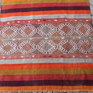 May include: A rectangular, hand-woven rug with a geometric pattern and a fringed edge. The rug features alternating bands of orange, red, and gray, with a central section of diamond and square motifs in shades of brown, white, and orange.