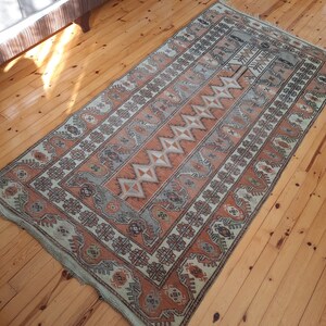 May include: A vintage rug with a geometric pattern in shades of beige, brown, and orange. The rug has a fringed edge and is laid out on a wooden floor.