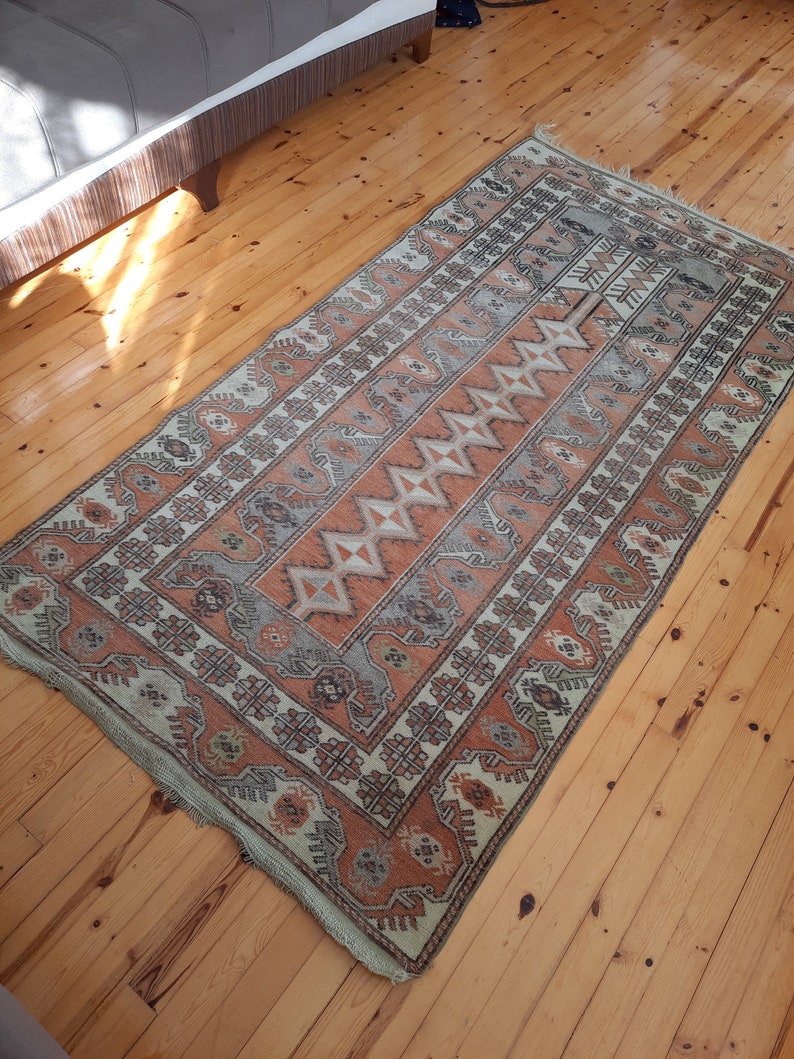 May include: A vintage rug with a geometric pattern in shades of beige, brown, and orange. The rug has a fringed edge and is laid out on a wooden floor.