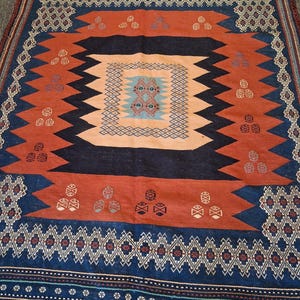 May include: A square, patterned rug with a central design in peach, surrounded by a dark blue and rust-colored border. The rug features geometric shapes and repeating patterns, with a diamond and zigzag design.
