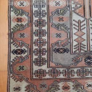 May include: A vintage rug with a cream background and intricate geometric patterns in shades of brown, pink, and green. The rug has a fringed edge.