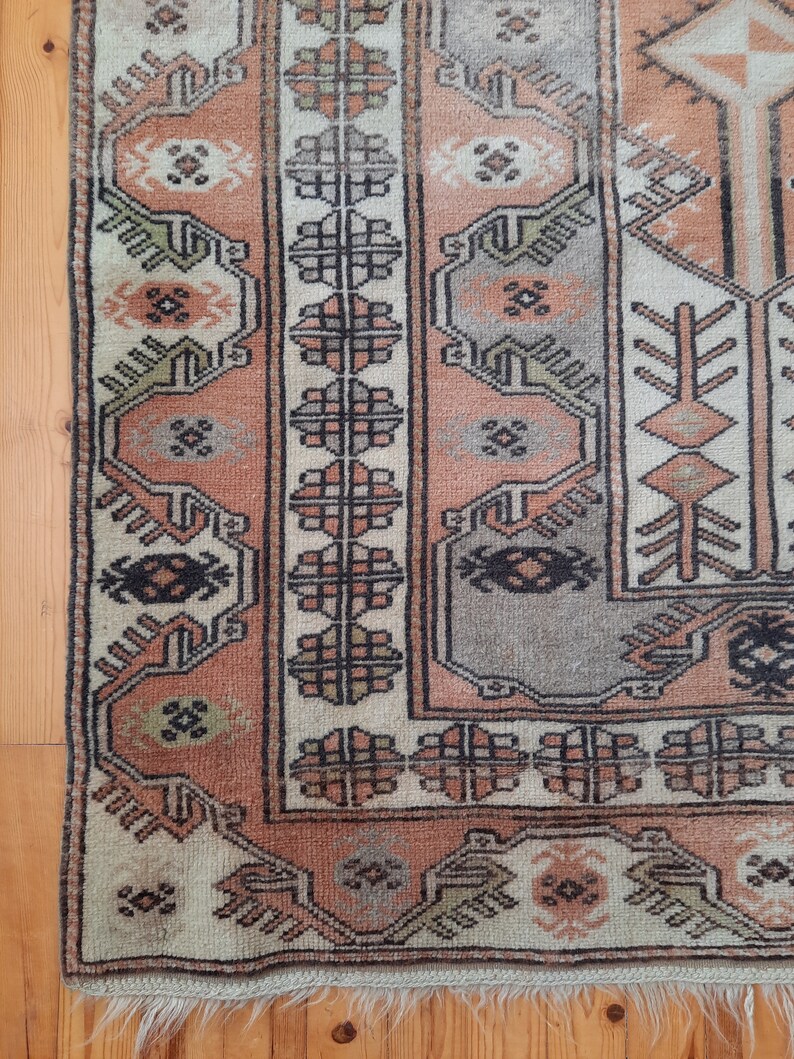 May include: A vintage rug with a cream background and intricate geometric patterns in shades of brown, pink, and green. The rug has a fringed edge.