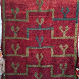 Peut inclure: Tapis rectangulaire tissé à la main, avec un fond rouge foncé et un motif géométrique répété en vert olive et sarcelle. Le motif présente des formes stylisées en forme d'arbre dans une bordure en escalier. Le tapis a une surface texturée et une bordure frangée.