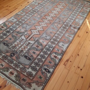 May include: A vintage rug with a geometric pattern in shades of beige, brown, and gray. The rug has a fringed edge and is laid out on a wooden floor.