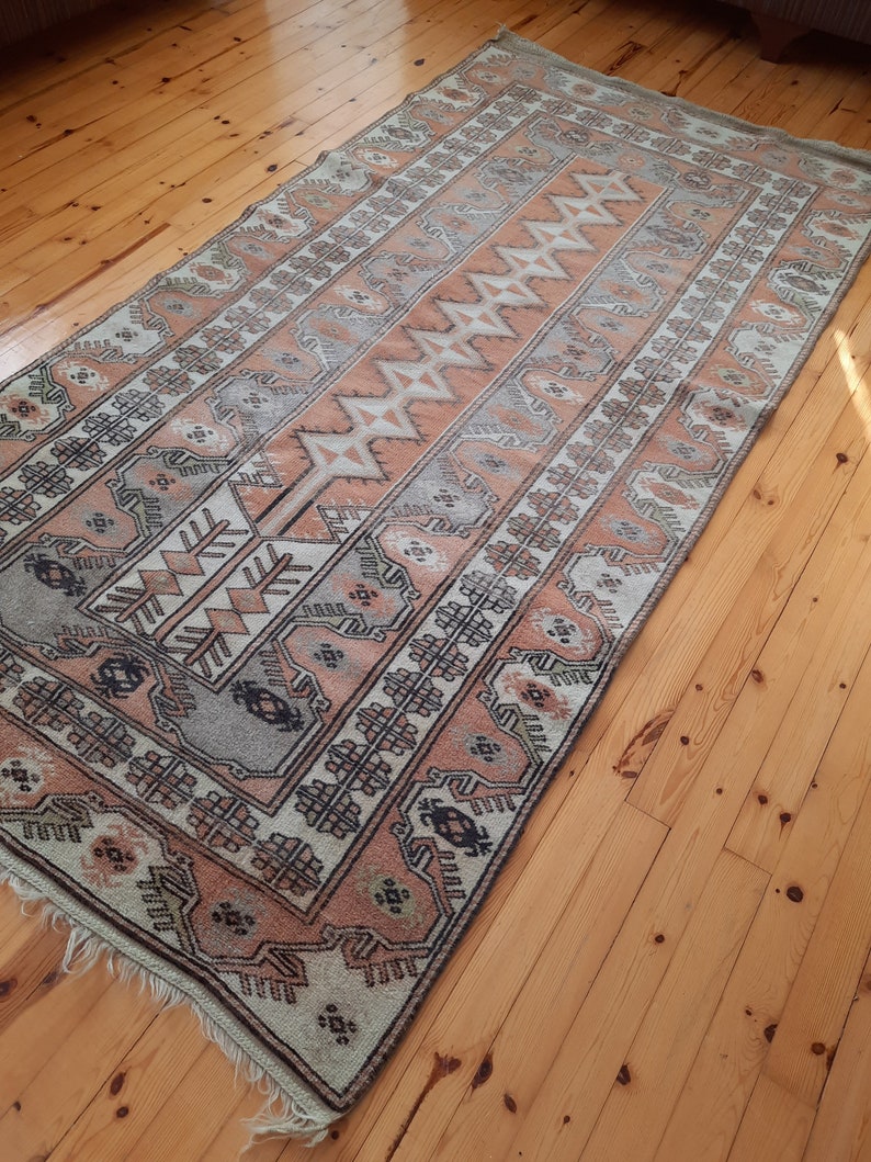 May include: A vintage rug with a geometric pattern in shades of beige, brown, and gray. The rug has a fringed edge and is laid out on a wooden floor.