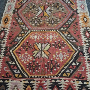 Puede incluir: Una alfombra rectangular tejida a mano con un diseño geométrico. La alfombra presenta un patrón central de color rojo, marrón y amarillo, rodeado de bordes negros y blancos. El diseño de la alfombra incluye formas de diamante y estrella.