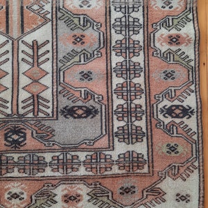 May include: A vintage rug with a traditional geometric pattern in shades of pink, beige, and black. The rug has a fringe border and is made of wool.