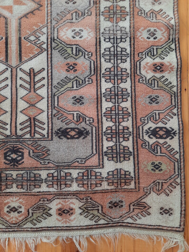 May include: A vintage rug with a traditional geometric pattern in shades of pink, beige, and black. The rug has a fringe border and is made of wool.