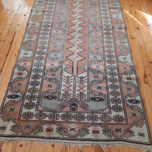 May include: A vintage rug with a traditional geometric pattern in shades of beige, brown, and pink. The rug has a fringed edge and is made of wool.