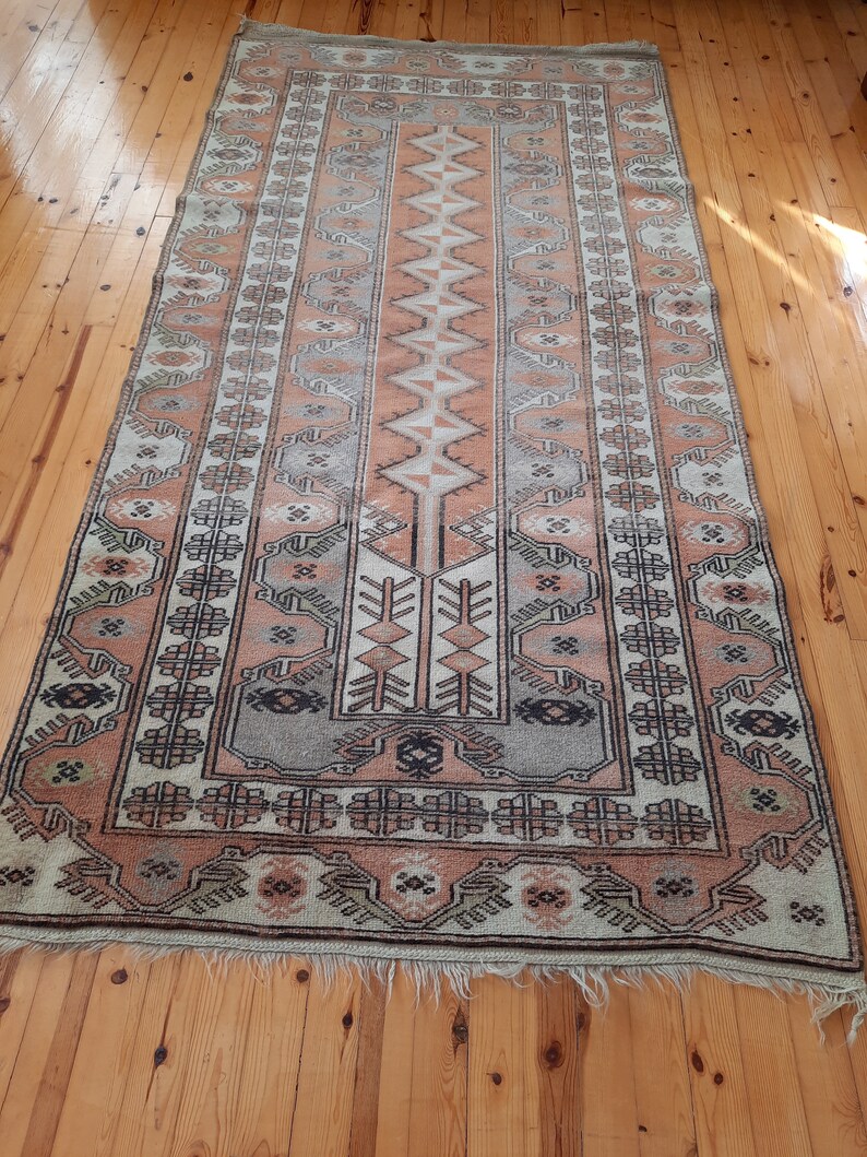 May include: A vintage rug with a traditional geometric pattern in shades of beige, brown, and pink. The rug has a fringed edge and is made of wool.