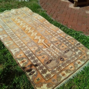 May include: A vintage, hand-woven rug with a geometric pattern in shades of beige, brown, and cream. The rug has a fringe border and is laid out on a grassy surface.