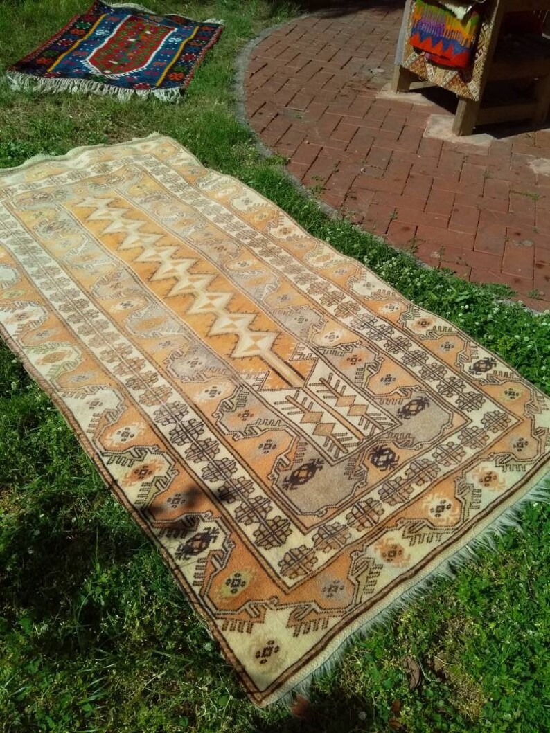 May include: A vintage, hand-woven rug with a geometric pattern in shades of beige, brown, and cream. The rug has a fringe border and is laid out on a grassy surface.