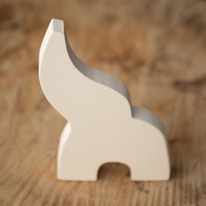 May include: A cream-colored, abstract wooden sculpture with a curved, upward-sweeping form. The sculpture has a rounded base with a cutout and a smooth, matte finish. The piece is set against a blurred wooden background.