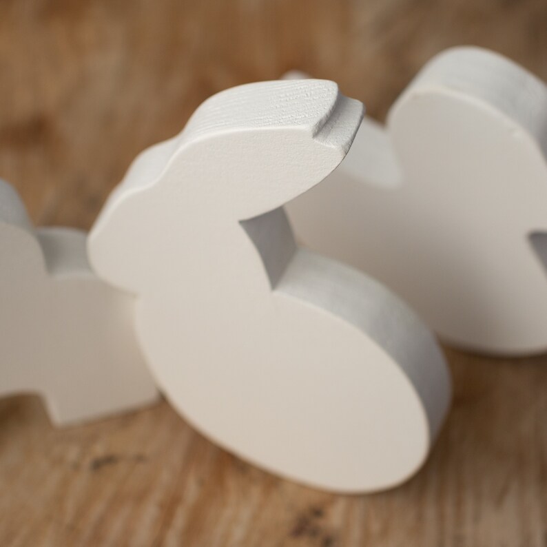 May include: Close-up of several white wooden bunny cutouts. The bunnies are in various stages of being cut out, with some still connected. The wood has a smooth, matte finish. The background is a light brown, wooden surface.
