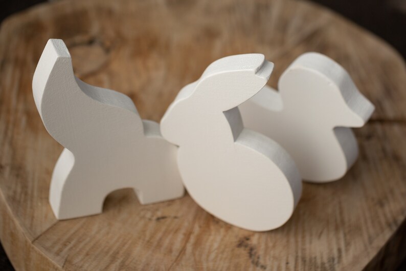 May include: Three white wooden animal figurines: a stylized elephant, a rabbit, and a duck. The figures are smooth and painted in a solid, neutral white color. They are arranged on a light brown, textured wooden surface.