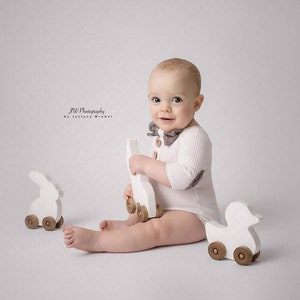 May include: A baby wearing a white ribbed bodysuit and a patterned bow tie, holding a white wooden toy. Two other wooden toys, a bunny and a duck, are nearby. The image is set against a plain white background.
