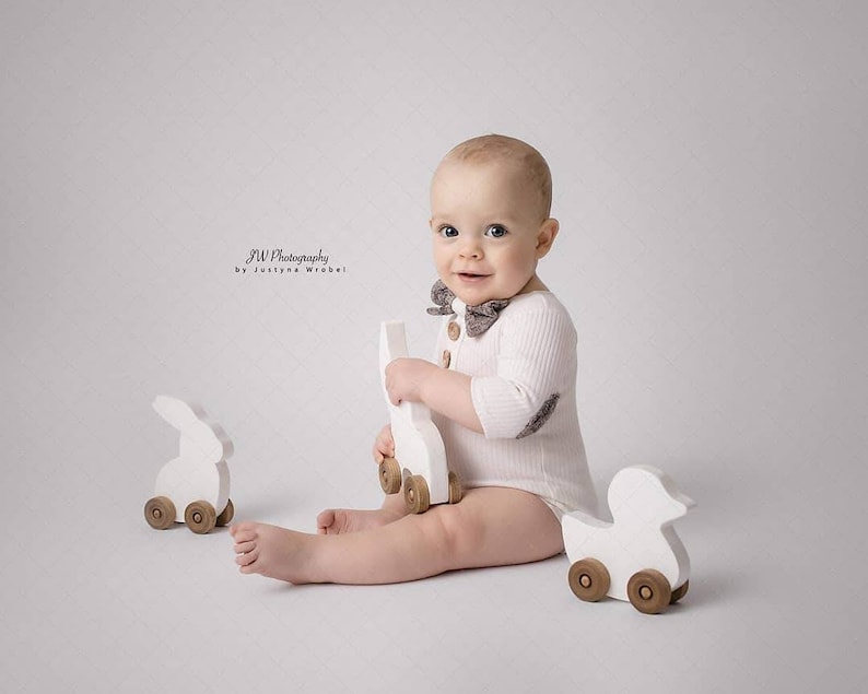 May include: A baby wearing a white ribbed bodysuit and a patterned bow tie, holding a white wooden toy. Two other wooden toys, a bunny and a duck, are nearby. The image is set against a plain white background.