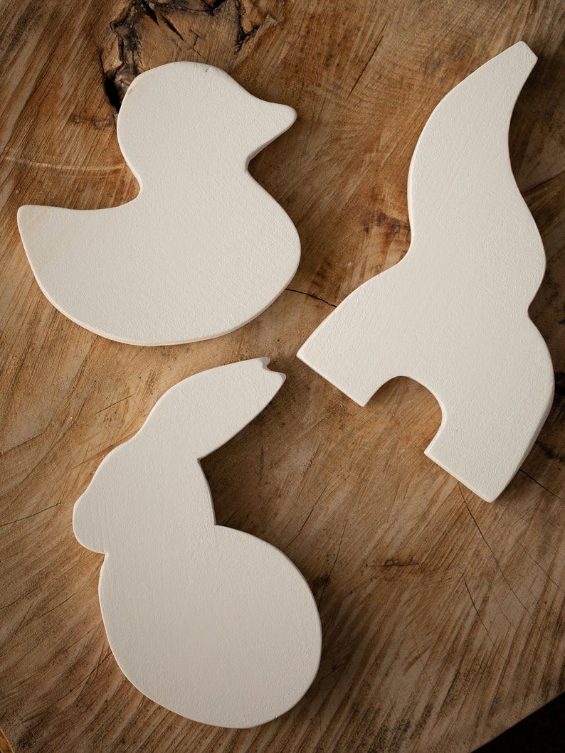 May include: Three off-white, wooden cutouts in the shapes of a duck, a bunny, and an abstract form. The duck is facing left, the bunny is facing right, and the abstract form is curved. The cutouts are on a textured, wooden surface.