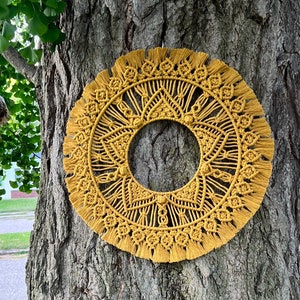 May include: A large, yellow macrame wall hanging with a circular design and fringe. The hanging is attached to a tree trunk.