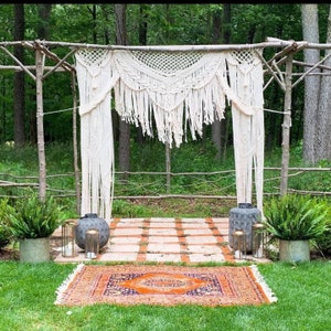 May include: A white macrame wedding archway with a woven design hangs from a wooden frame in a forest setting. The archway is decorated with two gray metal vases and a colorful rug is laid out on the ground in front of it.