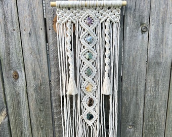 May include: A white macrame wall hanging with seven colorful gemstones woven into the design. The hanging is attached to a wooden dowel rod.