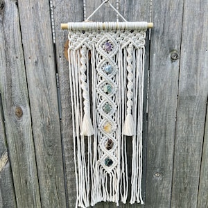 May include: A white macrame wall hanging with seven colourful gemstones woven into the design. The hanging is attached to a wooden dowel rod.