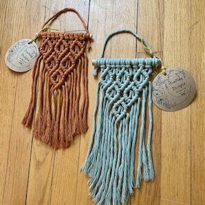May include: Two macrame wall hangings with wooden dowels. The one on the left is orange and the one on the right is light blue. Both have a diamond pattern and fringe. Each hanging has a circular tag with the text "Earthy Designs" and a floral design.