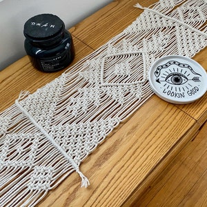 May include: A white macrame table runner with a diamond pattern. The runner is on a wooden table with a white ceramic plate with a black eye and the text "Lookin' Good".