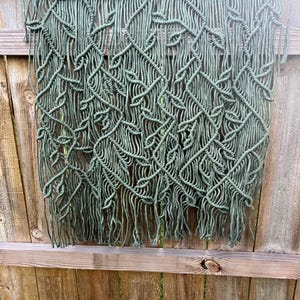 May include: A handmade macrame wall hanging in olive green. The intricate design features a woven pattern with a fringe at the bottom. The macrame is suspended from a natural wooden dowel, adding a touch of bohemian style to any space.