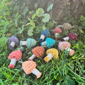 May include: A collection of colorful crocheted mushroom keychains with white accents and silver key rings. The keychains are arranged on a bed of green grass.