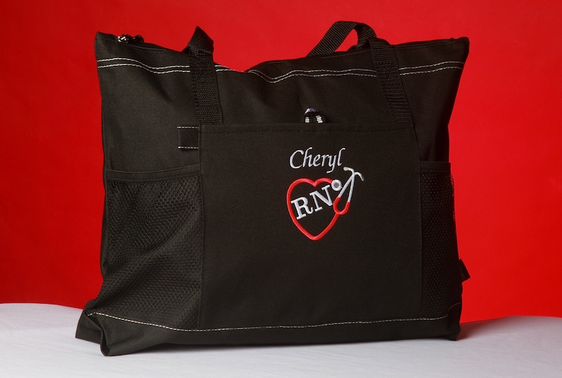 Peut inclure: Sac fourre-tout noir avec un c&oelig;ur rouge et les lettres "RN" brod&eacute;es dessus. Le nom "Cheryl" est brod&eacute; au-dessus du c&oelig;ur. Le sac a une poche en filet sur le c&ocirc;t&eacute;.