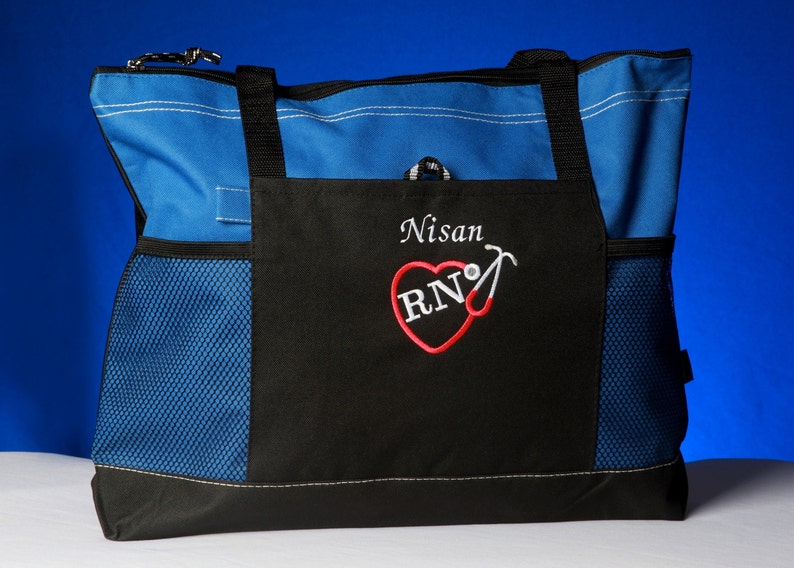 Peut inclure: Sac fourre-tout bleu et noir avec une poche en filet. Le sac est personnalis&eacute; avec un motif brod&eacute; avec le nom "Nisan" et un c&oelig;ur rouge avec les lettres "RN" et un st&eacute;thoscope.